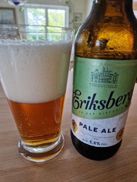 Eriksberg Pale Ale ölflaska och ett glas med skummande öl, inomhus, fönster i bakgrunden.