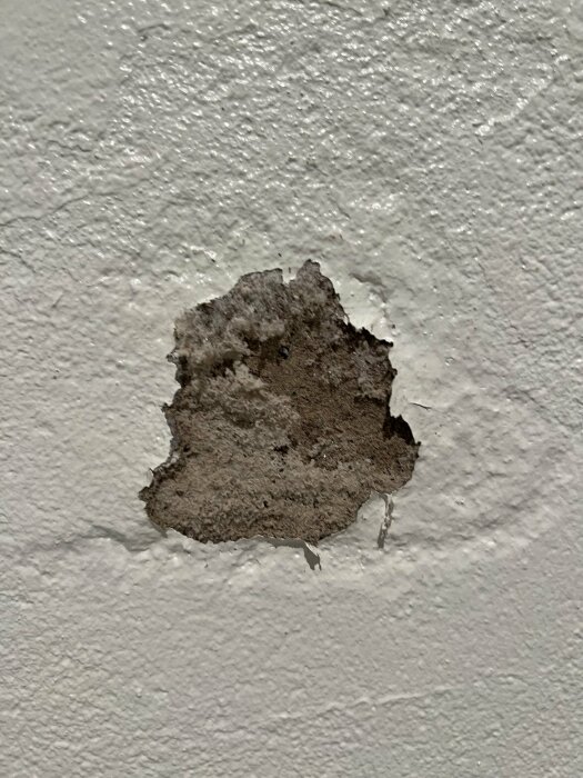 Ett skadat, avflagat område på en vit texturerad vägg som visar underliggande material.