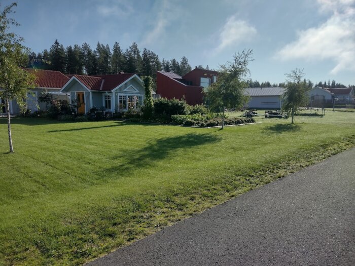 Ett välhållet enplanshus med trädgård, grönområde och röda uthus under solig himmel.