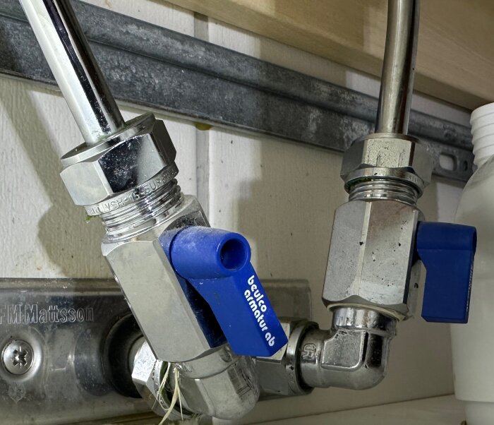 Vattenrörskopplingar och ventiler under en diskbänk, med blå markeringar och en etikett.