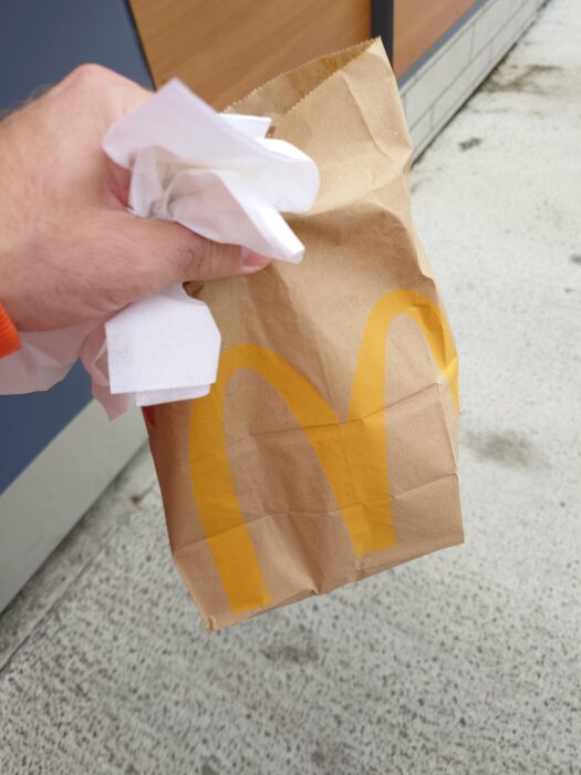 En hand håller en McDonald's papperspåse och servetter utomhus, med en suddig bakgrund av trottoar och snö.
