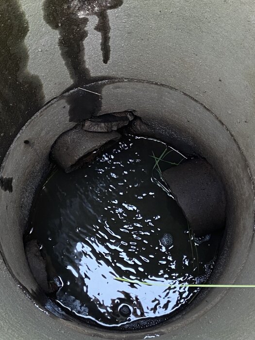 Rörformigt hål med vatten, skuggor och reflektioner, synlig betong och smuts, lite grönt växtstrå.