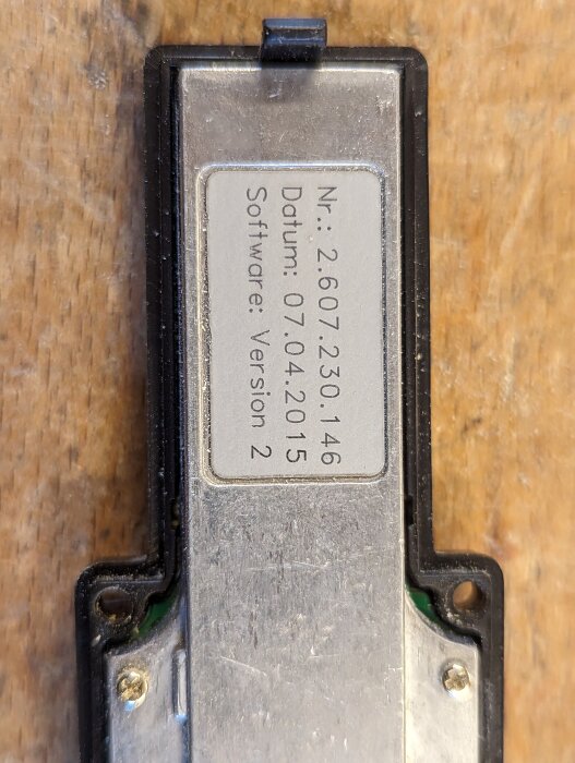 Elektronisk enhet med etikett som visar serienummer, datum och mjukvaruversion på träbakgrund.