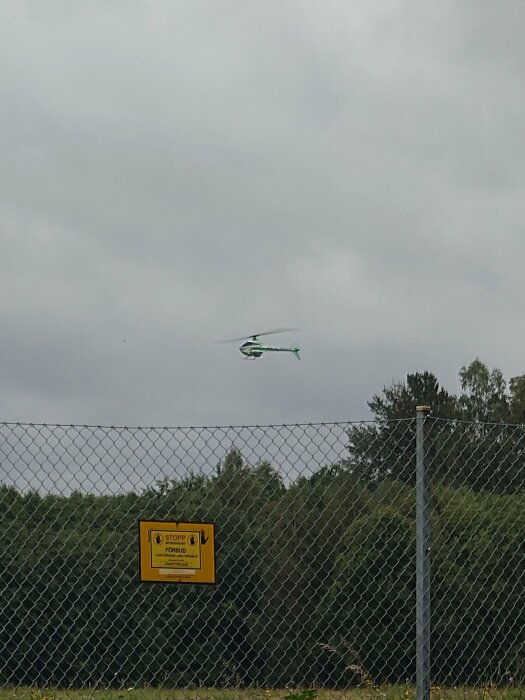 Helikopter flyger över staket med varningsskylt, mulet väder, grå himmel, träd i bakgrunden.