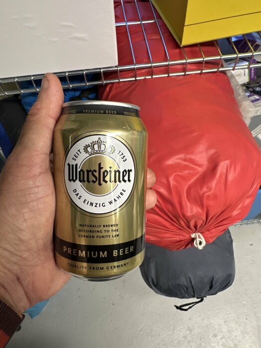 Hand håller Warsteiner ölburk, stående nära röd dyna och förvaringshyllor i rum.