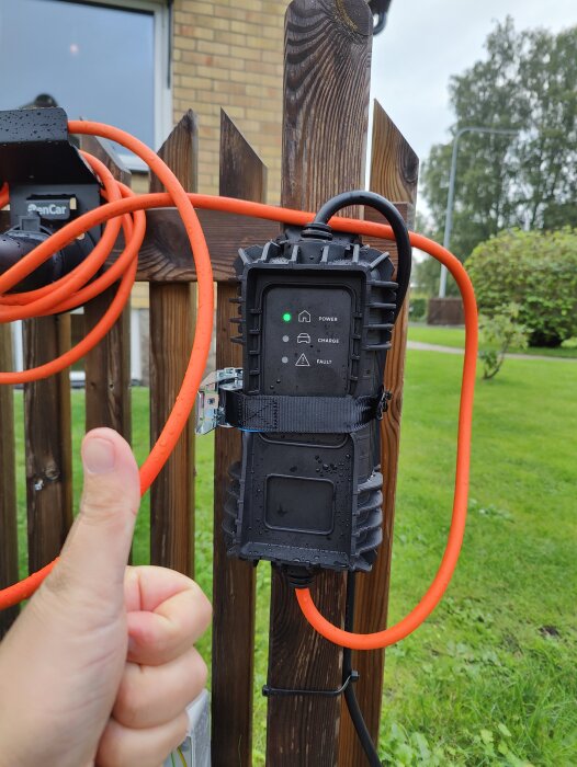 En hand ger tummen upp intill en svart, upphängd laddningsstation med orange kabel på trästaket.