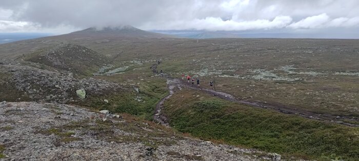 Vandrare på lerig stig i kargt fjällandskap under molnhimmel.