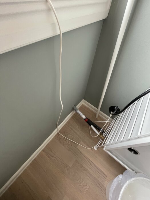 Hörn av rum med kablar längs väggen, golvstång, och sopkorgsdel.