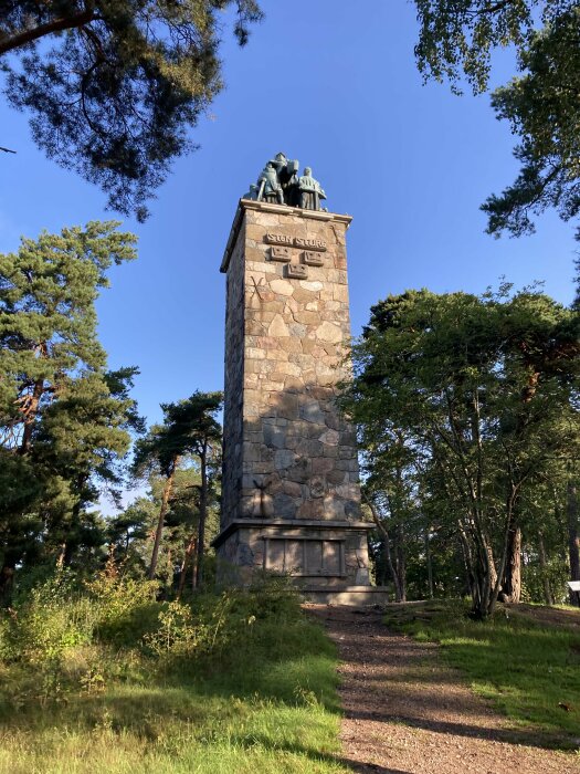 Stenpelare med skulpturgrupp på toppen, omgiven av träd och blå himmel, med text "Sven Stolpe".