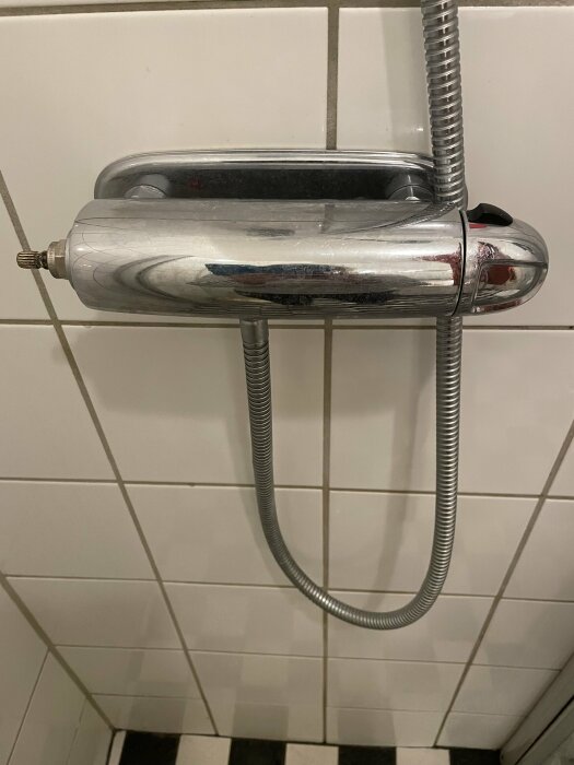 Väggmonterad handdusch och slang i badrum med vita kakelväggar.
