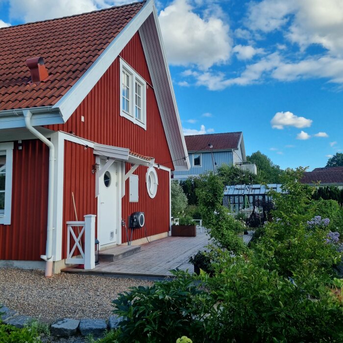 Rött trähus med vita knutar, veranda, grönskande trädgård och blå himmel.