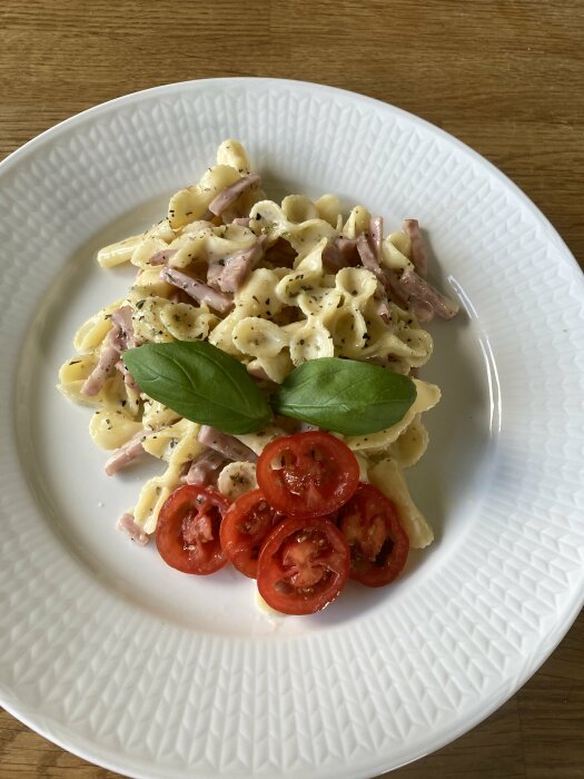 Ett fat med pasta, skinka, kryddor, färska tomat-skivor och basilikablad på träbord.