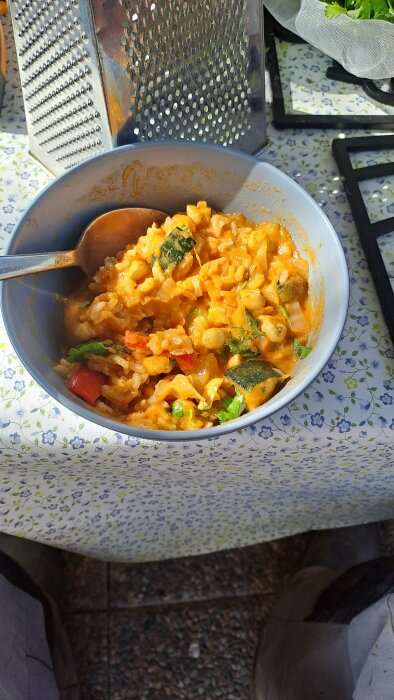 En skål med mat, möjligtvis pasta, grönsaker, utomhus på blommigt bord. Solsken, redskap, färsk persilja i bakgrunden.