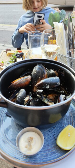 En person vid ett bord med mobil, framför en skål musslor, glas, citron och sås.