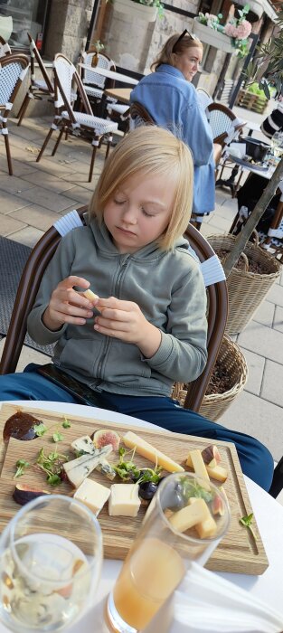 Barn sitter vid uteservering, tittar på mat, ostar på träbricka, avslappnad atmosfär, dagtid.