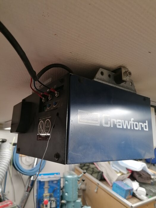 Blå Crawford box monterad under tak, elektriska kablar, verkstadsbakgrund, otydliga objekt i förgrunden.