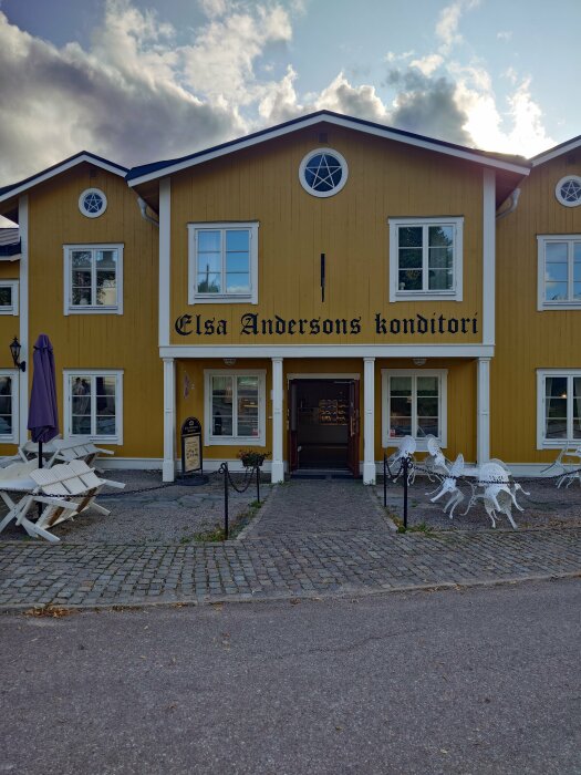 Gult tvåvånings konditori med text "Elsa Andersons konditori", fönster, vita utemöbler och kullersten, molnig himmel.