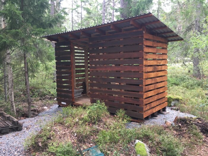 Träkonstruktion i skog, troligen vildmarks utedass eller vedbod, på grusbas, omgiven av gröna träd och vegetation.