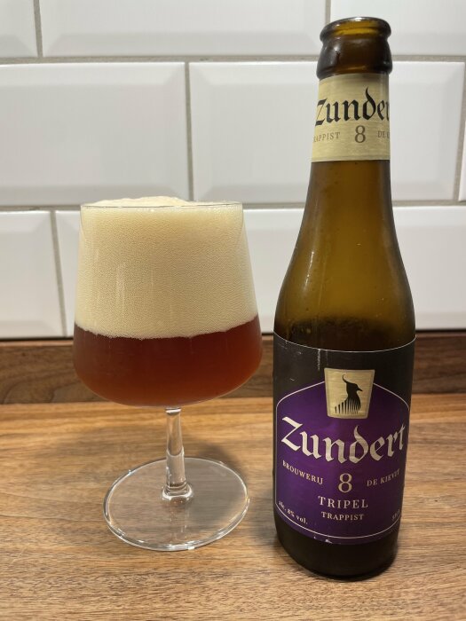 Flaska Zundert Trappist-öl bredvid glas med skummande öl, träyta, vita kakelväggar i bakgrunden.