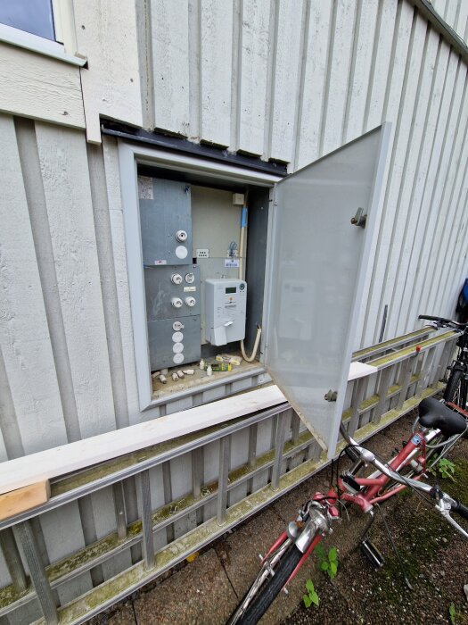 Ett elskåp öppet utanför byggnad med en röd cykel lutad mot staketet.