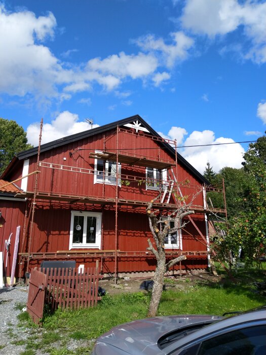 Rött hus med byggnadsställningar, blå himmel med moln, bil i förgrunden, träd bredvid huset.