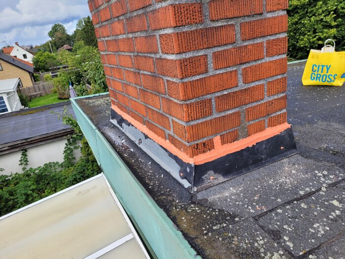 Röd tegelskorsten, mörkt tak, nymålat fundament, plastpåse, trädgårdar i bakgrunden, molnig himmel.