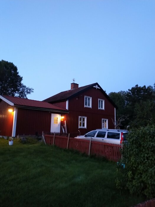 Ett rött trähus med vita fönsterkarmar vid skymning, upplyst av yttre lampor, staket och grönt gräs.