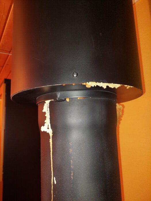 Svart metallpelare med rost och skumrester på orange bakgrund. Möjlig skadedjursbekämpning eller läckage.