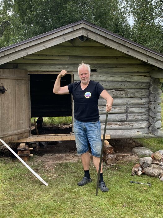 Man poserar med spade framför timrad bod, visar musklerna, grönska bakom, stickerbild på tröja, somrig dag.