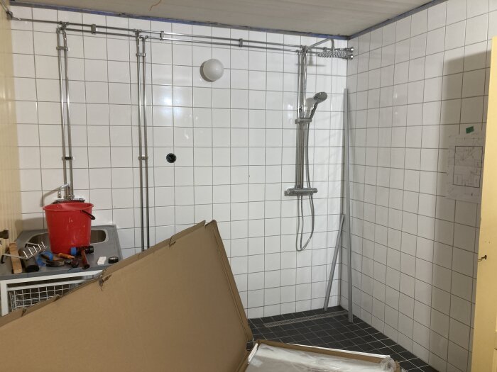 Ett rörigt duschrum med verktyg och en kartong. Renovering pågår möjligen. Vit kakelvägg och svart golv.