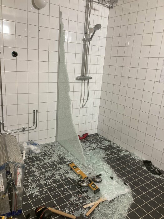 Ett badrum under renovering med krossad glasdörr, verktyg och spridda byggmaterial på golvet.