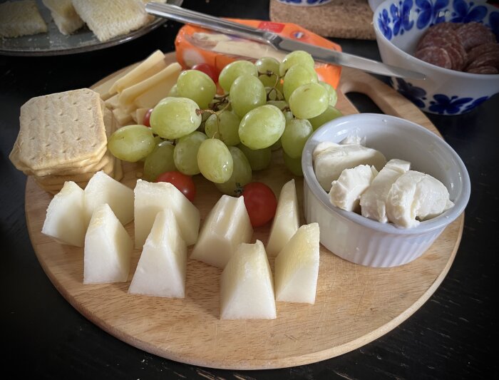 Ett ostbricka med gröna druvor, kex, tomater, melon och portionsostar på ett träfat.
