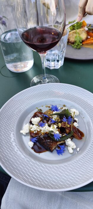 Elegant middagsinställning med vin, vattenglas och en estetisk tallrik med gourmetmat, dekorerad med blommor och örter.