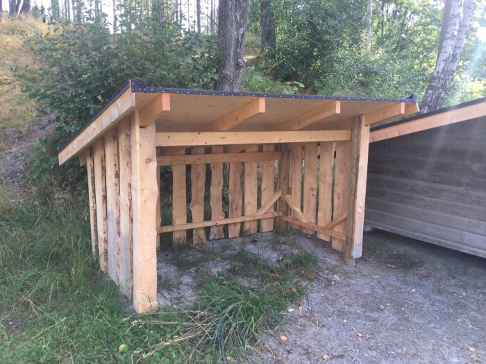 Ett nybyggt träskyddsrum eller vedskjul i en skogsmiljö med en äldre konstruktion intill.