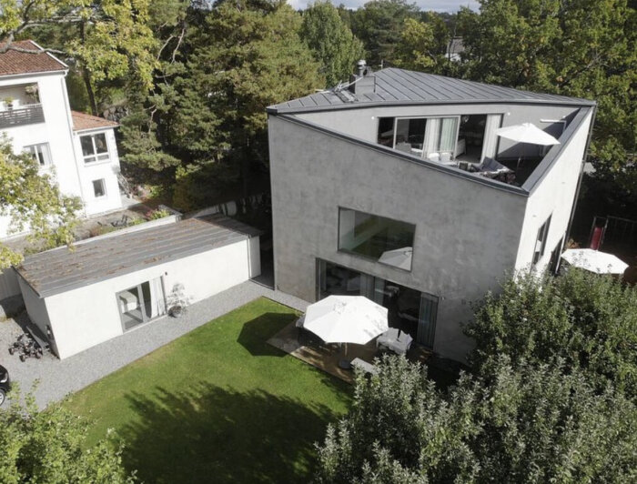 Modernt hus med slät fasad, gräsmatta, paraplyer, terrass och omgivande grönska, sett från luften.