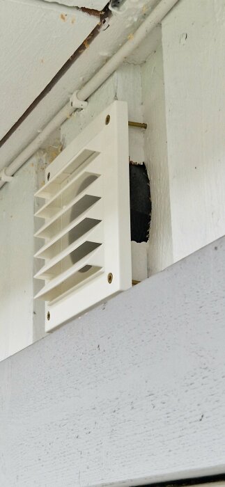 Vit ventilationsgaller på vägg med synligt rör och ojämnt hål bakom galler.