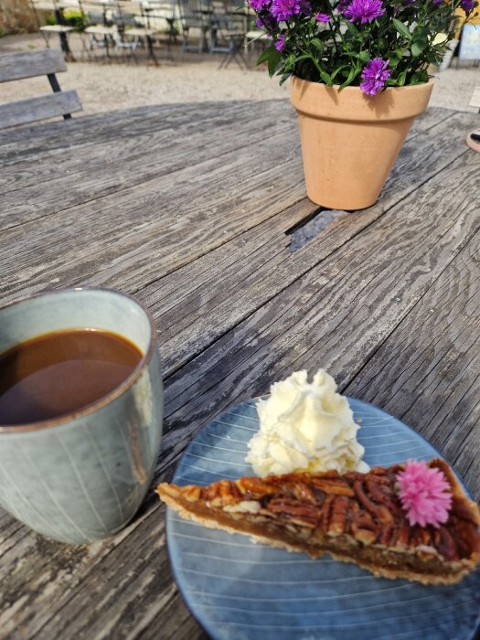 Kaffe och pecannötspaj med vispgrädde på blå tallrik vid träbord utomhus. Blomkruka i bakgrunden.