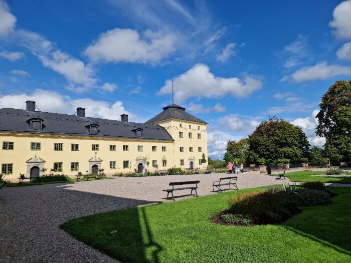 Gult slott, grusgång, gröna trädgårdar, parkbänkar, blå himmel med moln, besökare.