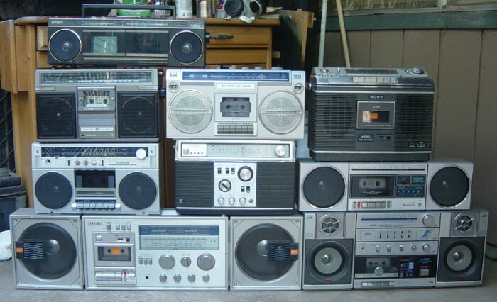 Samling av vintage boomboxes, varierande storlek och märken, framför ett garage. Retro musikspelare.