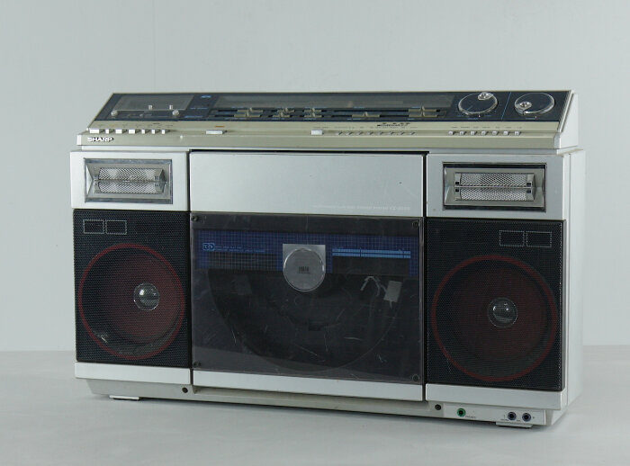 Vintage, silverfärgad boombox med kassettspelare, högtalare, knappar och rattar på en vit bakgrund.