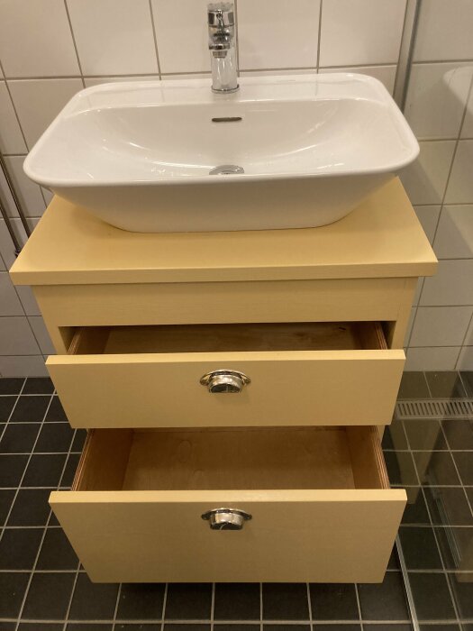 Vit handfat ovanpå beige förvaringsmöbler med öppna lådor, kakelvägg, mörka golvplattor.