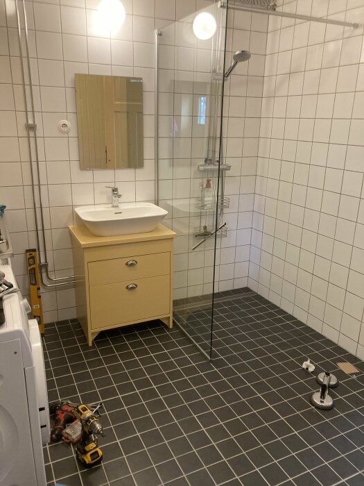 Badrum, vit kakel, duschhörna, beige handfatsskåp, spegel, verktyg på golvet, tvättmaskin, renoveringsarbete pågår.