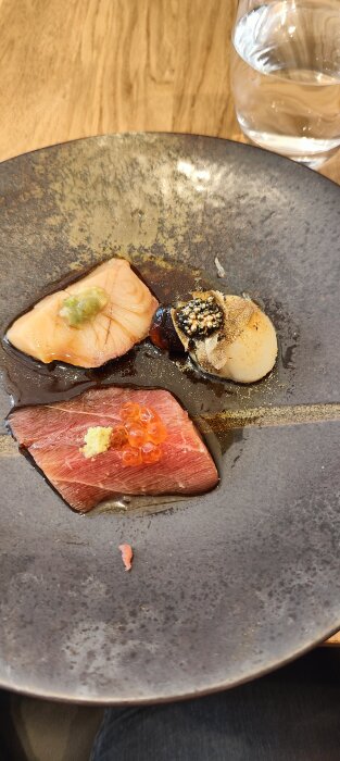 Förrättstallrik med sushi-ingredienser, lax, tonfisk, kammussla, kaviar, vatten i bakgrunden, modern presentation.