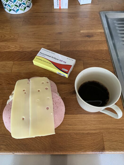 Ost- och skinkmacka med kaffe, medicinförpackningar på ett träbord. Morgonstart eller fikapaus, hemmiljö.