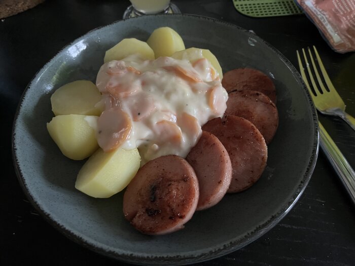 En måltid med potatis, skivad stekt falukorv och sås på en mörk tallrik. Enkelt och vardagligt.