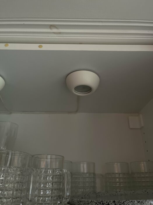 Inbyggt takljus med genomskinliga dricksglas på hylla under. Gråmålat skåp med beslag och ljusgrå vägg.