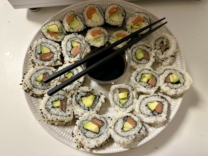 Flera sushi-rullar med lax och avokado på ett vitt fat, inklusive pinnar och sojasås.
