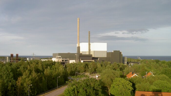 Industribyggnad med skorstenar, grönska runtomkring, havet i bakgrunden, moln på himmelen, klar dagtid.