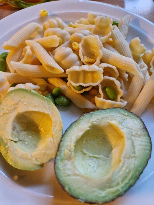 Ett måltid med pasta, gröna ärtor och en halverad avokado på en vit tallrik.
