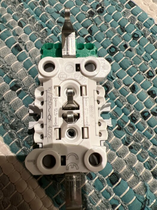 Elektrisk installationskomponent, troligen en strömbrytare, mot textil bakgrund. Monteringsdetaljer synliga. Fokus på teknisk utrustning.